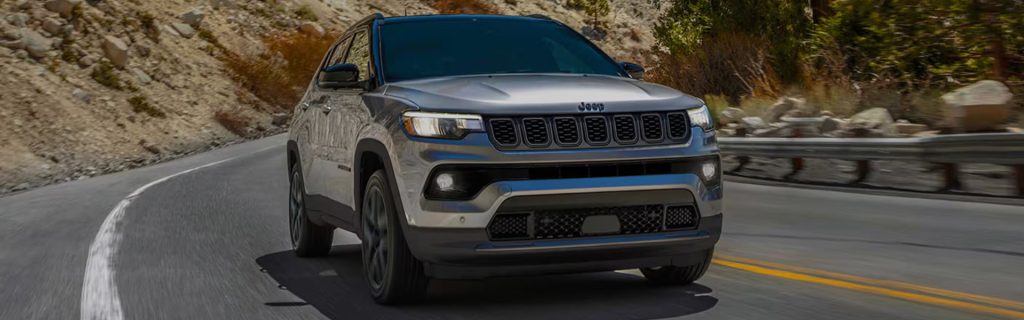 A 2026 Jeep Compass navigates a winding mountain road surrounded by trees and rocky terrain.