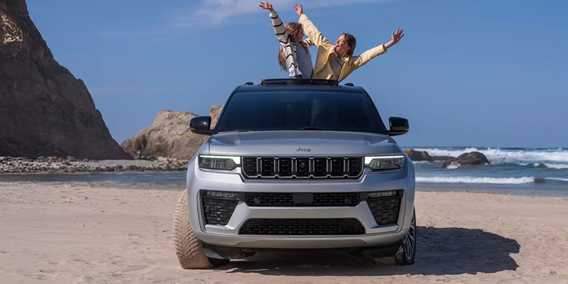 The 2026 Jeep Grand Cherokee on the beach with a man and woman sitting on the hood, relaxing and taking in the seaside scenery.