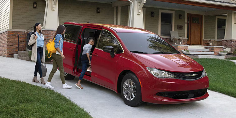 A red Chrysler vehicle parked in front of a house, surrounded by people enjoying the outdoors.
