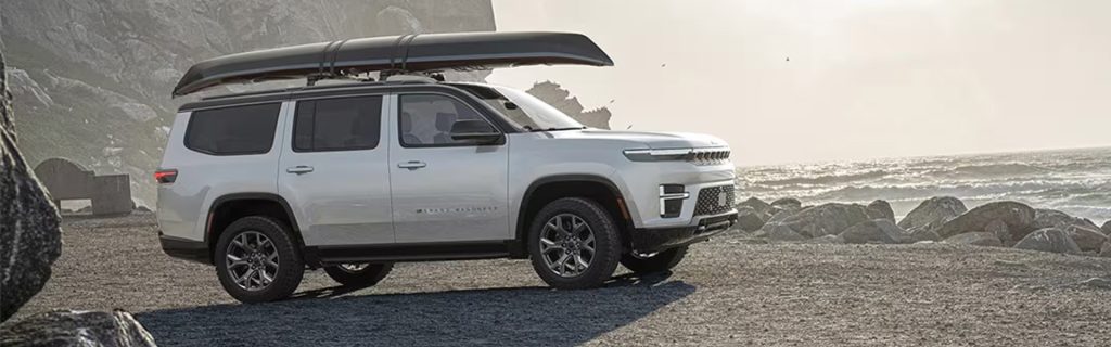 White Jeep SUV parked near a rocky beach with a rooftop cargo carrier and ocean view in the background.