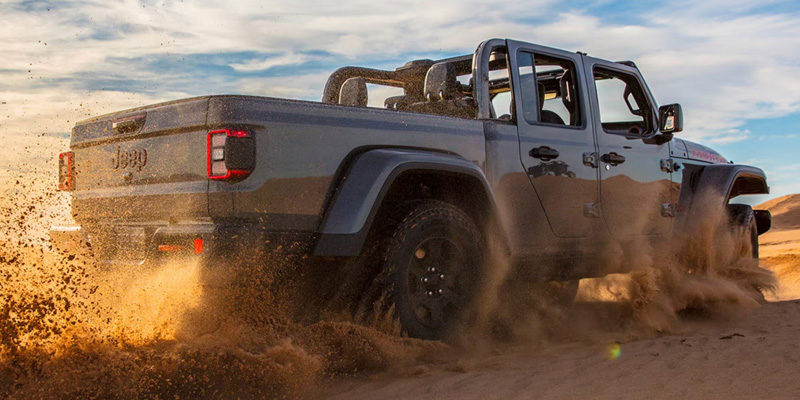 A 2026 Jeep Gladiator truck driving through sandy terrain, kicking up dust and sand.
