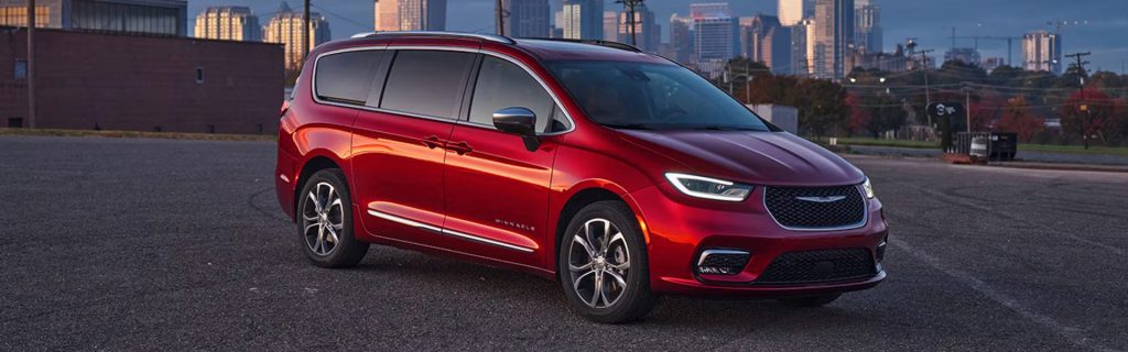 A red Chrysler Pacifica parked in an urban setting with a city skyline in the background during sunset.