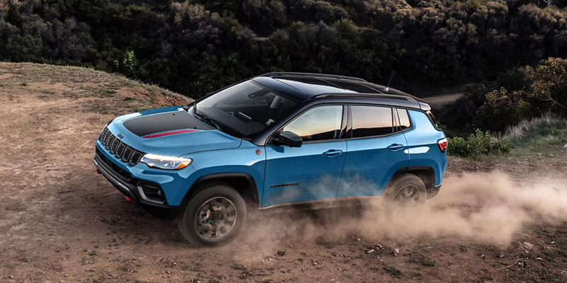 A blue Jeep Compass SUV driving on a dirt path in a rugged terrain, kicking up dust.