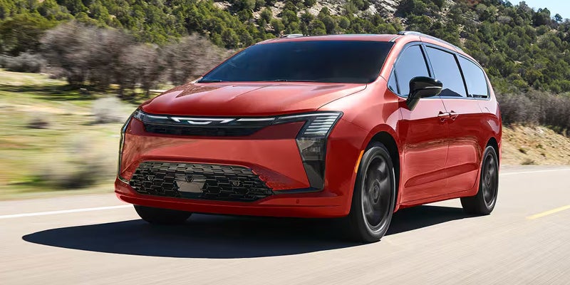 A red Chrysler Pacifica driving on a road with green hills in the background.