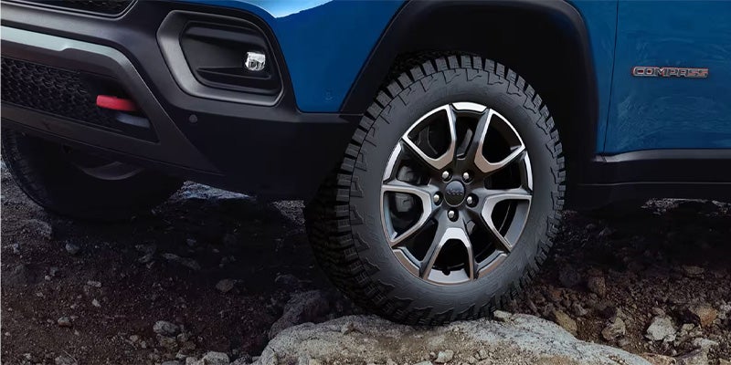Close-up of a blue Jeep Compass front tire on rugged rocky terrain, showcasing off-road capability and detailed alloy wheel design.