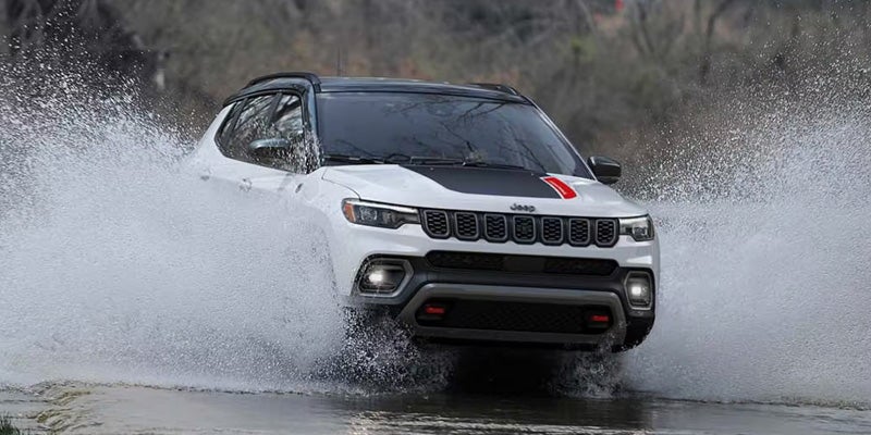 White Jeep Compass SUV splashing through water on a muddy off-road trail with trees in the background.