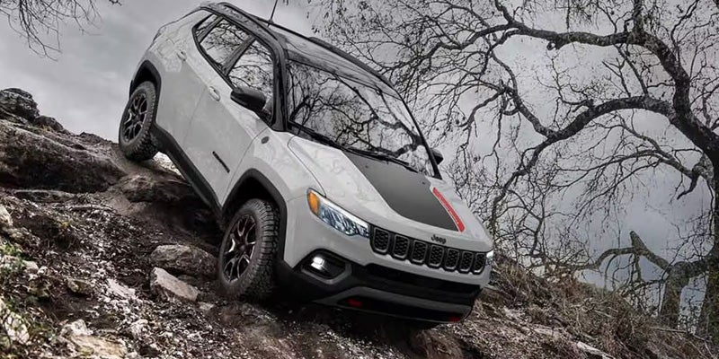 A white Jeep Compass parked on rocky terrain with a dramatic, overcast sky and leafless trees in the background.