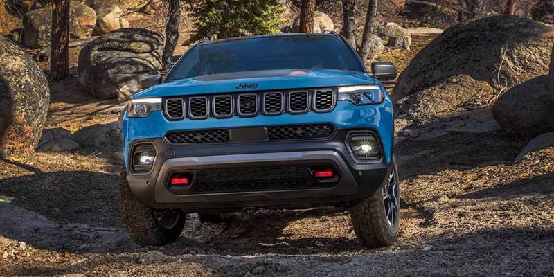 Blue Jeep Compass navigating rocky off-road terrain surrounded by large boulders and trees in a forested area.