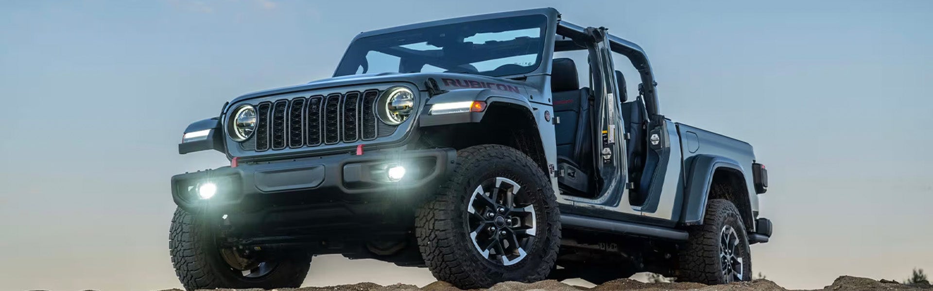 A blue Jeep Wrangler with all doors removed parked on a sandy terrain under a clear sky.