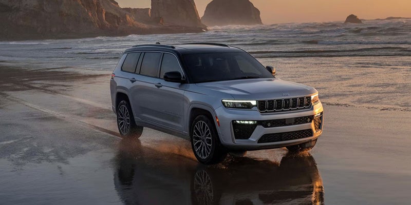 A silver Jeep Grand Cherokee parked on a beach during sunset, with waves in the background and rocky cliffs on the side.