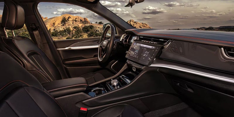 Interior view of a Jeep Grand Cherokee showcasing the dashboard, steering wheel, and seats against a backdrop of a scenic landscape with clouds and hills.