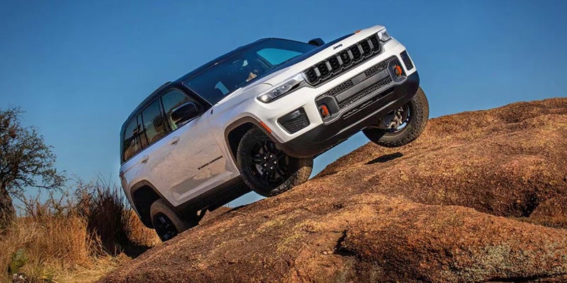 A white 2026 Jeep Grand Cherokee is driving up a rocky terrain against a clear blue sky.