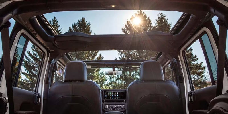 Interior view of a Jeep Wrangler with a panoramic sunroof, showcasing the front seats and dashboard, with trees and sunlight visible outside.