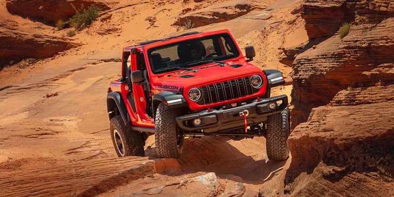 A red Jeep Wrangler Rubicon climbing over rocky terrain in a desert landscape.