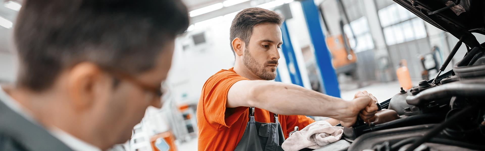 A mechanic working on a car engine while another person observes.