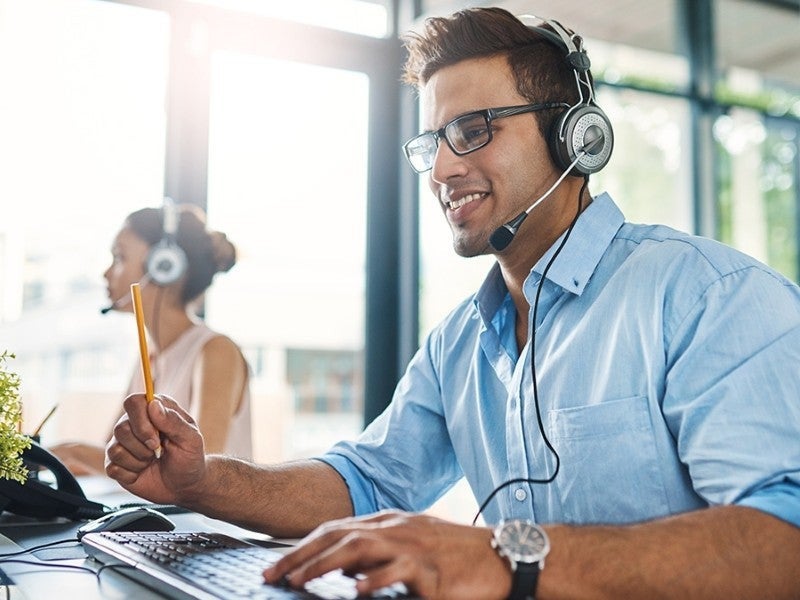 Customer service representative smiling, wearing headset, typing on keyboard in a bright office.
