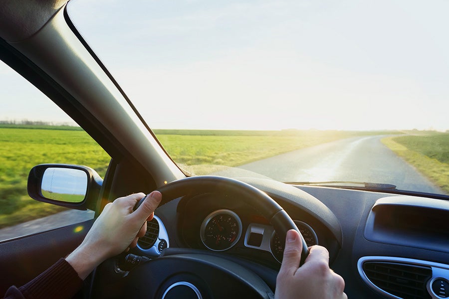 Person driving a car on a sunlit rural road surrounded by green fields.