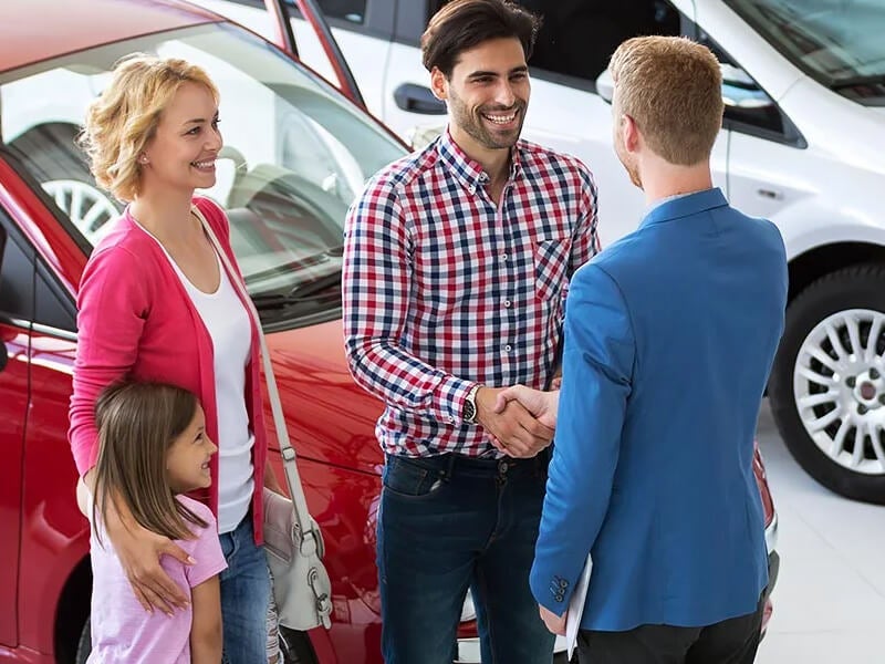 Family finalizes car purchase, shaking hands with a salesman at dealership.