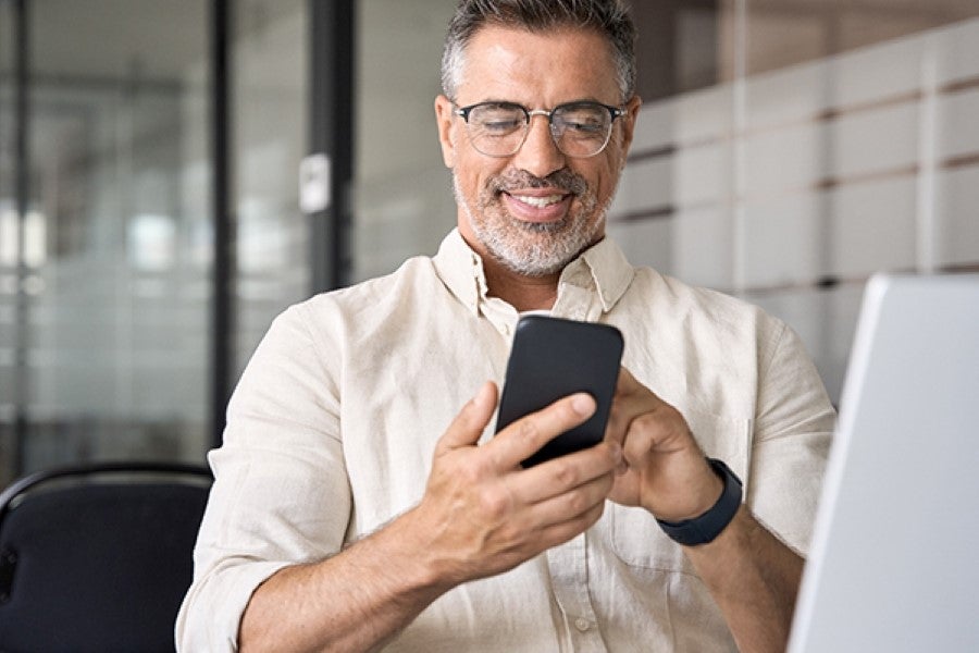 Smiling middle-aged man using smartphone, sitting at desk with laptop in modern office.