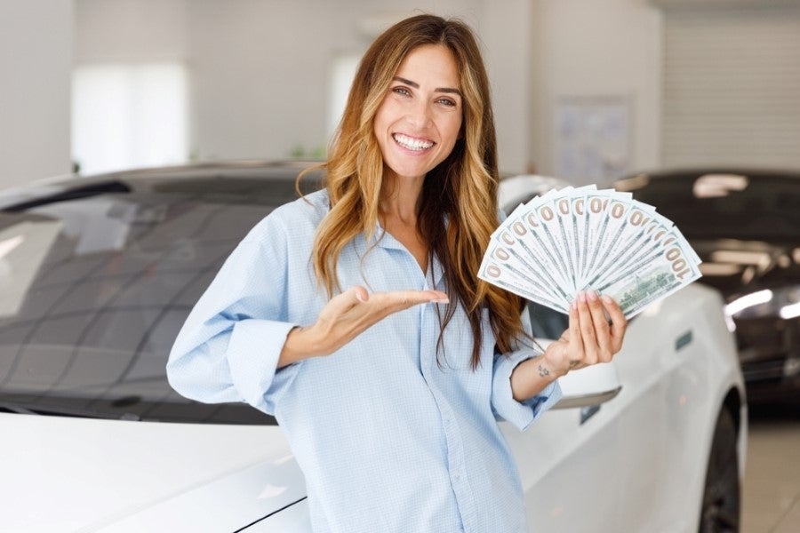 Happy woman showing cash in front of a white car at a dealership.