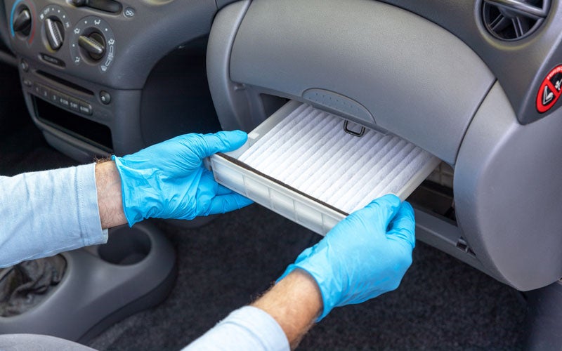 A person wearing blue gloves is replacing a cabin air filter in a car's glove compartment.